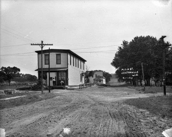 View down unpaved road towards an intersection on Main Street. On the left is a two-story wood frame building with two men and a dog on the porch and posters in the windows. A horse and buggy is parked in front. Alert Hall is further down the road on the left. On the right are trees and a flyer announcing a fair posted on a power pole.