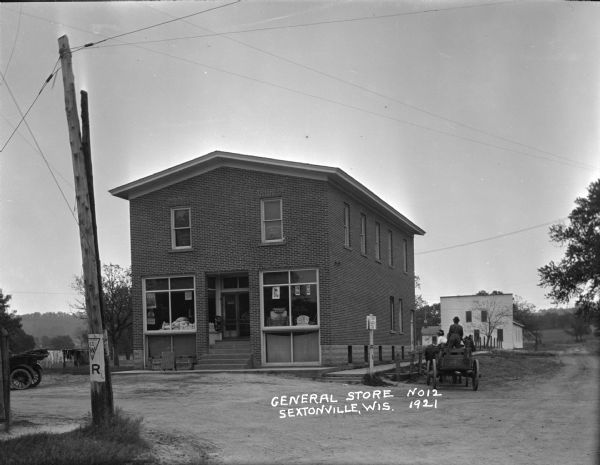 View across street of the Sextonville General Store, a two-story brick building on a corner. A car is parked on the left, and two men are sitting in a horse-drawn wagon on the right. Laundry is hanging out to dry in the background on the left. The Alert Hall building is in the far background.