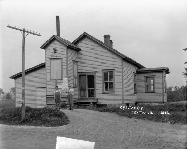 Exterior view of the Creamery, a small wood frame building. Two workmen wearing overalls are standing on the drive in front. Behind them next to the entrance is a loading area with a set of doors at wagon height, and a block and tackle with a hook attached near the roof line. Broadsides advertising silos are posted below the doors. There is a box attached to the power line in the foreground.