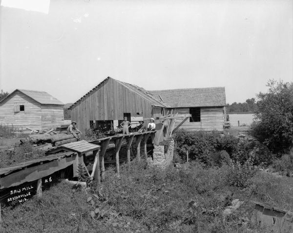A sawmill with workmen sitting outdoors in front of the mill. One man sits on a stack of logs, the other three men stand on a roof which is open sided and protects the belt-driven engine. A river or lake is in the background.