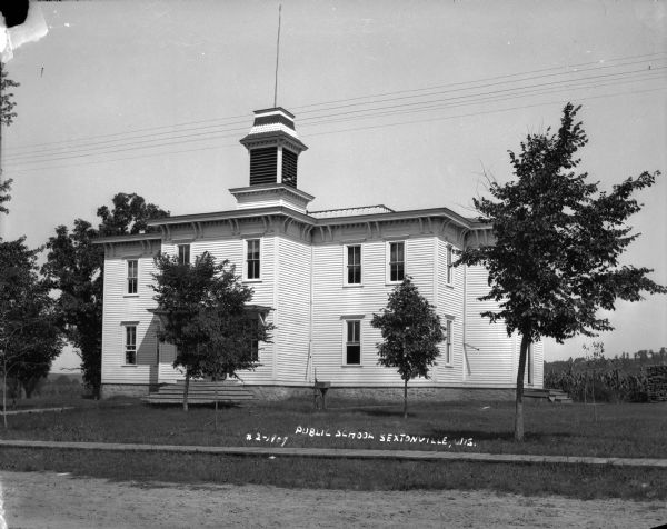 Exterior view of the public school from unpaved road. The building is a two-story wood frame building with a flat roof and bell tower with a flag pole on top. There is a hand-pump near the steps leading to the entrance. Trees are in the front yard. Behind the school is a field, and on the far right a stack of fuelwood.