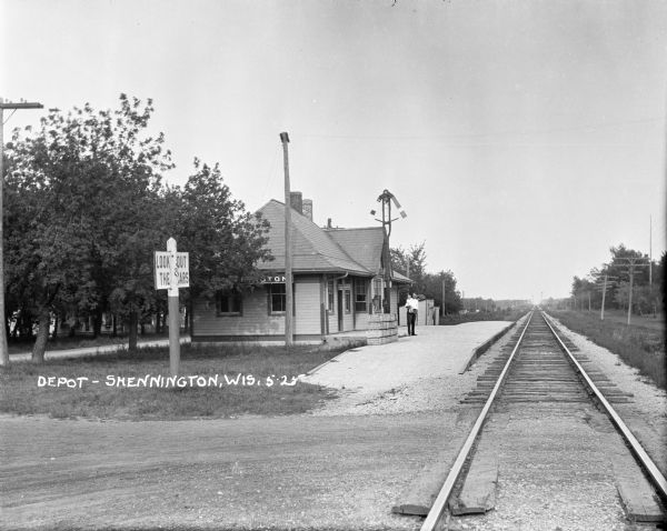View down railroad tracks and across road towards the depot. A sign posted at the crossing says: Look Out for the Cars." A man with a small child is standing on the platform near stacks of crates. A sign for Western Union telegraph is posted next to the door.