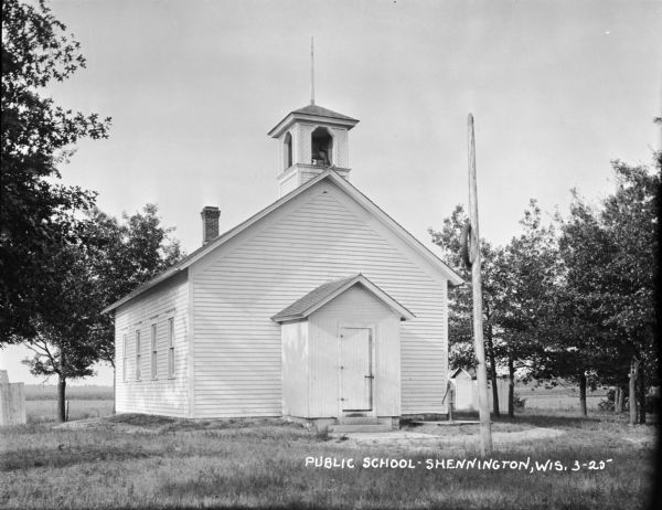 A one-story wood frame school with a lightning rod or flagpole on the bell tower. A hand water pump is near the front entrance, and a tire swing is wrapped high around a pole in the yard directly in front of the entrance. Outhouses are behind the school on the left and right, and cornfields are in the distance.