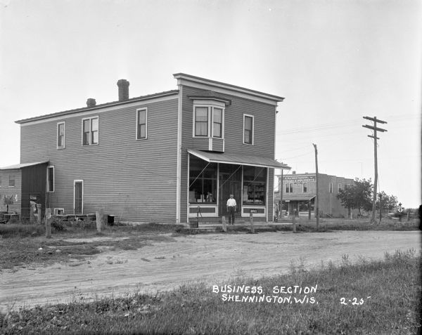 View across an unpaved road of the business district. There is a man standing in front of a general store, with a hand water pump and bucket near the porch, and a dog sitting by a screen door in the side yard on the left. In the background is a man standing in front of a hardware store/post office with a gas pump.