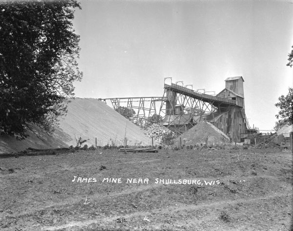 Exterior view of James Mine with a field and fence in the foreground. Tram tracks lead from the tall building to large piles of rocks. There are steep stairs up to the entrance.