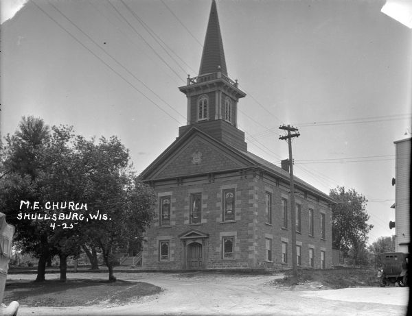 Exterior view of the Methodist Episcopal Church, a stone building with stained glass windows, a belfry, and a steeple. There is a house next door in the background on the left. Cars are parked in the foreground, and a tall commercial building can be partially seen on the right.