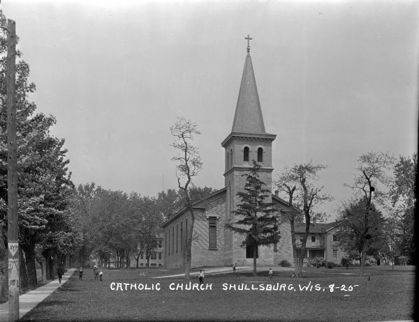 View across lawn of exterior of the Catholic Church, a stone and brick building with stained glass windows. A man is walking down the tree-lined sidewalk on the left, and children are playing in the church yard. Trees, houses and buildings are in the background.
