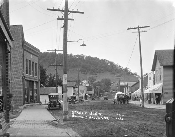 View down sidewalk of (Hwy 30 W) with commercial buildings on both sides. A steep hill is in the background. On the left are parked cars near storefronts in brick buildings. Behind a parked car two women can be seen sitting outside a small millinery, and a shoe shop is further down the street. Two horses pull a heavily loaded wagon on the right near more storefronts.