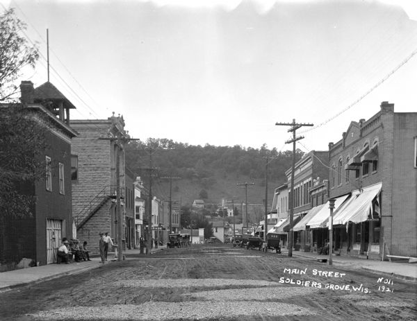 View down center of Main Street. On the left a group of men are gathered on the sidewalk near a drinking fountain which is in front of a brick building with a bell tower. A barber and a service garage are further down. On the right is a pool hall and restaurant, and other unidentified storefronts. Automobiles are parked along the curb.