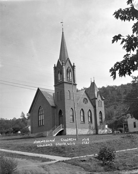 View from unpaved road of the front and right sides of the Norwegian Church, which is a brick building with arched stained glass windows, a belfry and a steeple with a weather vane. Below the large arched stained glass window in the front is a bulkhead in the stone foundation. Behind the church on the right is a house. In the foreground is a sidewalk. In the background is a steep hill with trees.