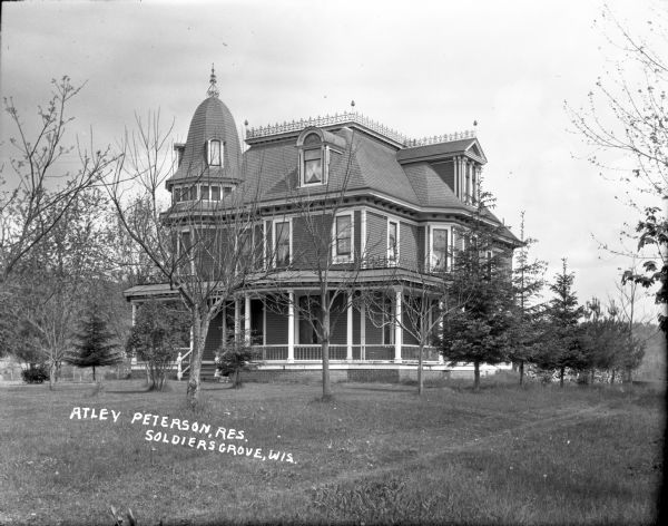 Exterior view across lawn of the Atley Peterson home, which is a two-story Victorian with an attic and a turret. Metal work is along the flat section of the roof. There is a wrap-around porch and lace curtains in the windows. Pine trees and bushes are planted in the yard. A large pile of fuelwood is behind the house on the right.