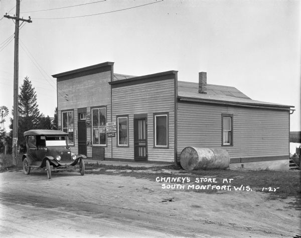 View across road towards Chaney's Cash General Store. Behind trees on the left is a house and windmill. Laundry is drying behind the store on the right. A car is parked in front of the store, which has a number of advertisements for tobacco, flour and cookies near the front windows.