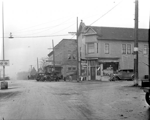 View down unpaved street in a central business district. A Rexall Drug Store is on the corner on the right. A large stone building, which has a sign for "HO_B's Service Garage," is next door further down the street with numerous cars parked in front. On the left is a "Tourist Camp" sign on a pole set in the middle of the street on a concrete base which says "Keep To Right."