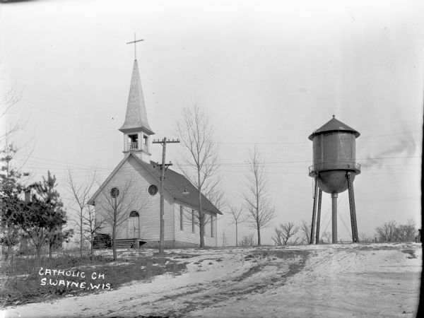 View up a road towards the front of the catholic church, which has a steeple and belfry. The double door entrance has an arched stained glass window, and above that are two round stained glass windows. A water tower on the right, and another building can be partially seen behind trees on the left.