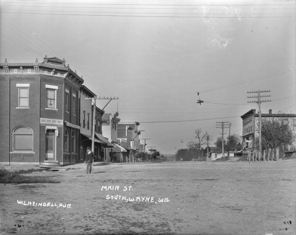 View down Main Street. On the left a bank is on the corner with a Masonic symbol in the upstairs window. A general store is down the block on the left. Horses and buggies are hitched to the posts near the sidewalk. Street lights are suspended over the street. Pedestrians are in the street and along the sidewalk.