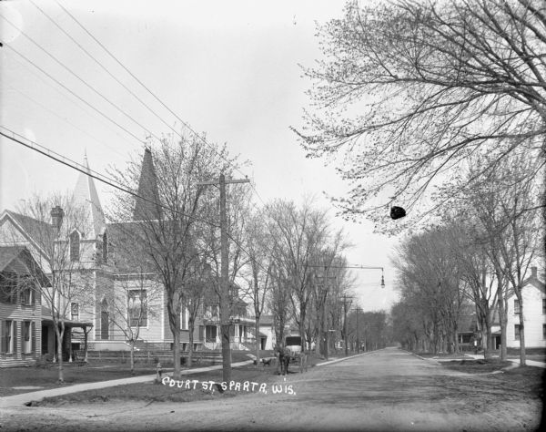 View from street of the Court Street neighborhood. On the left are a church and houses. There is a fire hydrant in the foreground, and nearby parked near the curb is a horse and buggy. Two dogs are near a tree on the terrace. Houses line the street on the right. A street light is suspended above the street.
