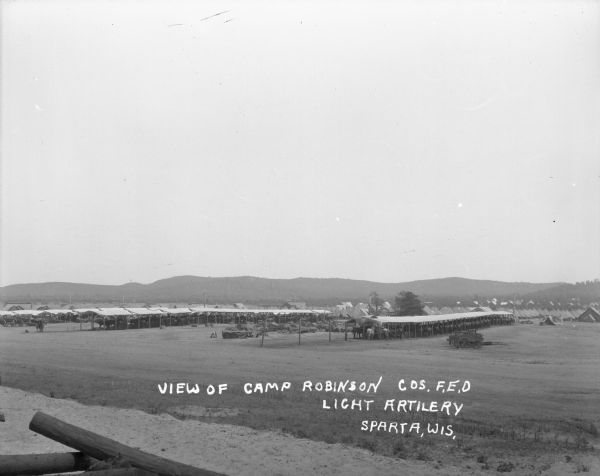 View of Camp Robinson towards an open area with cannons grouped together in the center between rows of horses under shelters. There are wagons on the right. Tents, barracks and buildings are behind the horses, and there are hills or bluffs in the far distance.