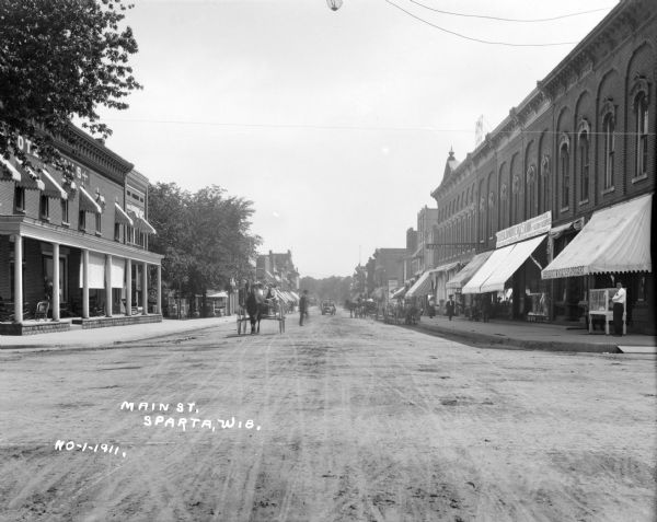 View down center of Main Street. On the left is the Hotel Lewis, with a columned porch and awnings above the second-story windows. Further down the street is a cemetery, a bowling alley and a restaurant. On the right side of the street is a grocer and a mercantile. Horses and buggies are traveling on the street, and pedestrians are walking along the sidewalk.