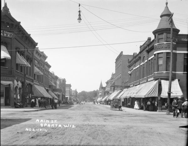 View of Main Street from its intersection with Oak Street. On the left corner is a bank and two drug stores. On the right are two restaurants and a shoe shop. Most of the storefronts and windows have striped awnings. Horses and buggies and automobiles are in the street. Many pedestrians are along the sidewalks and a street light is suspended over the intersection.