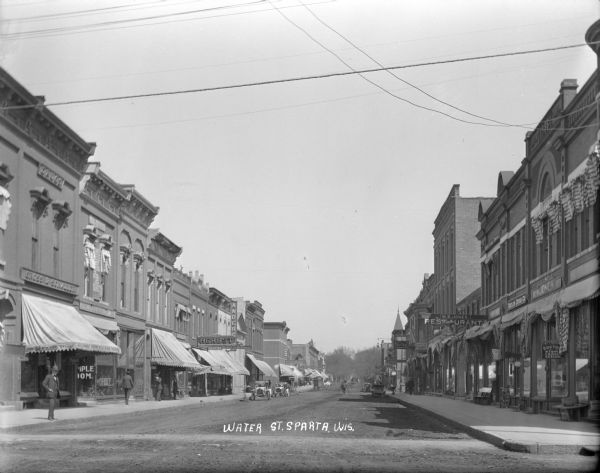 View down Water Street. The buildings on the left side of the street have their awnings down, and the buildings on the right, which is in shade, have them folded up. A drug store and dentist's office are on the left. A doctor's office, restaurant and a shoe shop are on the right. Automobiles are parked along the curb, and horse-drawn vehicles are in the distance. Bicycles are leaning against storefronts, and  pedestrians walk along the sidewalk.