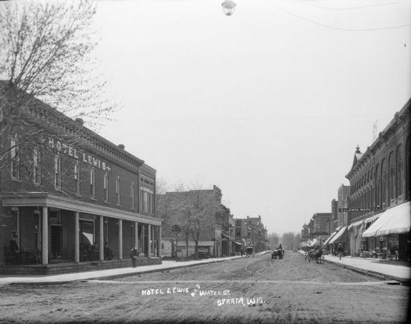View from street of the Hotel Lewis and other buildings on along the left. The hotel has a large front porch, with men relaxing in rocking chairs. A mail carrier stands on the sidewalk in front of the hotel. On the right side of the street is a Mercantile and other storefronts. Horse-drawn vehicles are in the street. A street light is suspended over the intersection.