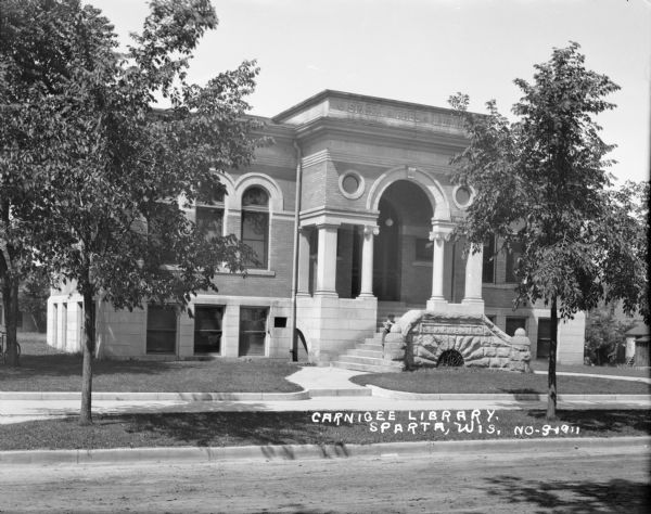 View from street of the Carnegie Library. A boy is standing on the stone steps that lead up to the arched entrance which has two round windows and a datestone of 1902. Along the top of the building just below the roof line is carved: "Sparta Free Library". Caption reads: "Carnigee[sic] Library, Sparta, Wis."