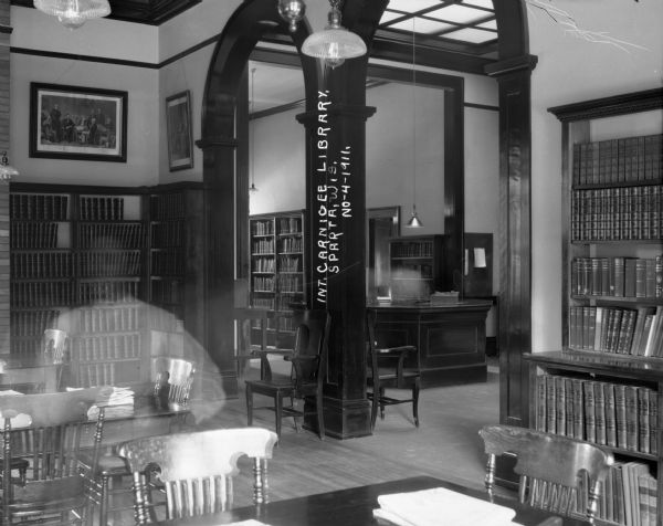 A view of the interior of the Carnegie Library. There is a faint image of a woman in the left foreground. Bookshelves line the walls, tables and chairs are nearby, and a large archway leads to a front desk. There are lithographs above the bookshelves on the walls. Electric lamps hang from the ceiling, and a large skylight is above the front desk.