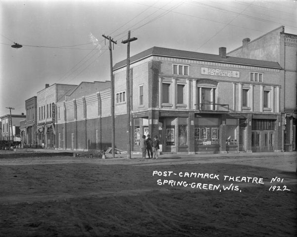 View from unpaved street of the Post-Cammack Theatre on a corner, with coming attraction posters in the windows. Three men are standing on the sidewalk in front. A bowling alley and a tire shop are behind the theater on the left. A barber shop is next door on the right.