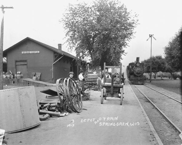 View down side of railroad tracks towards the Spring Green depot, with a locomotive just pulling in. A group of passengers are standing on the platform. In the foreground are two men and two young boys who stand near a cart that holds baggage. There are piles of pipe, carriage wheels and other items stacked near the railroad tracks. Other people sitting under the trees across the tracks.