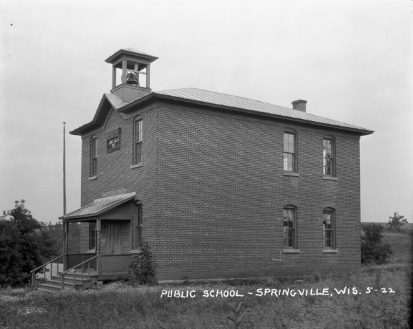 Exterior view of the public school, a two-story brick building in a rural area with small bell tower on the roof above the entrance. There is a flagpole on the side of the roofed porch over the entrance. A sign between two windows on the second floor reads: "Joint Dist No 1." The school yard has long, unmowed grass, and there are fields and trees in the background.
