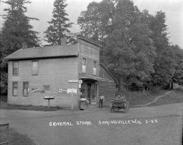 View from road of the general store, a two-story wood frame building in a rural area. A man and a child are standing near the door. Another child is sitting in a parked car. A sign pointing to Hwy H is on the wall with other posters. A number of mailboxes are mounted on a wheel sitting on top of a wooden pole. On the right is a path leading up into trees.