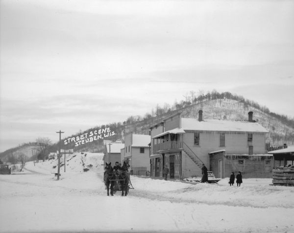 View from road of a winter street scene. A man is driving a horse and wagon in the road. Two men and two children stand on the sidewalk. A person can be seen standing behind a second-story window of a storefront, and there is a row of businesses on the right. In the background is tree-covered hill.