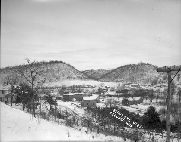 View from hill down towards homes and businesses surrounded by more hills. A bridge over the Kickapoo River is in the center. Railroad cars are parked on the tracks near the bridge.