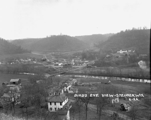 View from hill looking down at homes and businesses in town. A bridge goes over the Kickapoo River. The depot and railroad tracks and Main Street are behind it. The public school is against the hill on the right.