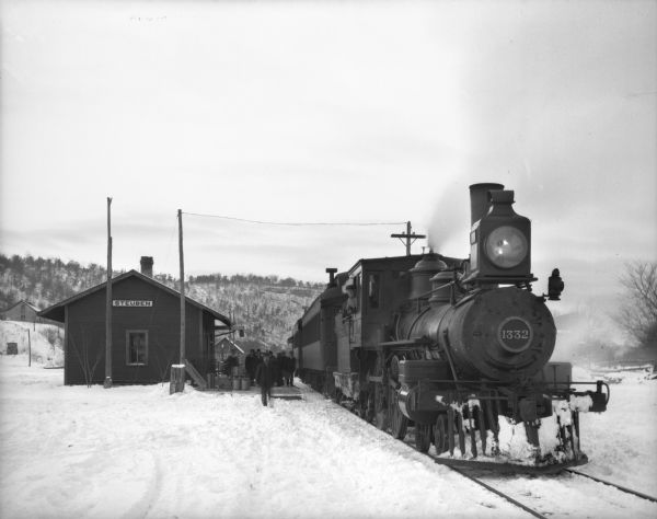 A winter view of the depot with a passenger train. Passengers are on the platform. The engineer is leaning out the window of the locomotive. In the background is a tree-lined ridge.