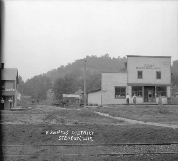 View across railroad tracks of the business district. On the left two people stand in front of a building on the corner with a signs that says: "Office". Across the road to the right a man and two women stand in front of a general store. In the background are steep, tree-covered hills.