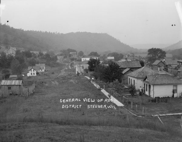 View from a hill towards houses and outbuildings. A man stands on a board sidewalk in front of a group of houses. A woman and young girl stand near the entrance to one of the houses. In the background an unpaved street winds along towards other buildings.Wire fences separating the properties. In the far background is a long, tree-lined ridge and a valley.