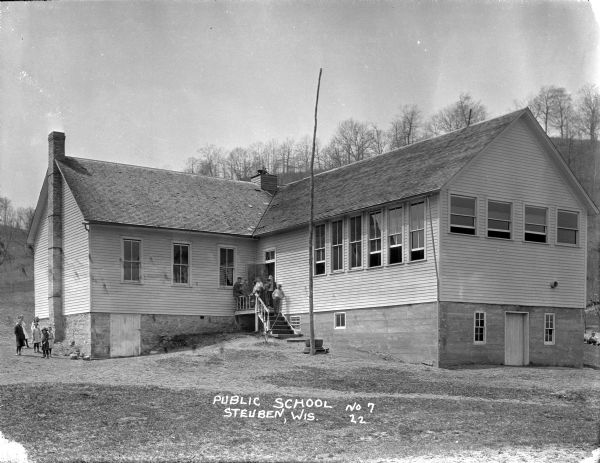 View across yard of the public school, a one-story wood frame L-shaped building on a hill. There are groups of students standing on the stairs at the entrance, on the left side of the building near the stone foundation and chimney, and lounging on the lawn behind the school building on the right. A small bell is on the roof in the center of the building.