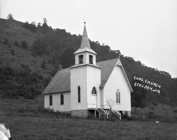 Three-quarter view of the front and side of the Congregational Church at the bottom of a steep, tree-lined hill. A wooden building with narrow arched windows, belfry and steeple. On the top of the steeple is a lightning rod with a weather vane. Near the foot of the steps is a new tree supported by a wooden post, and a barrel.