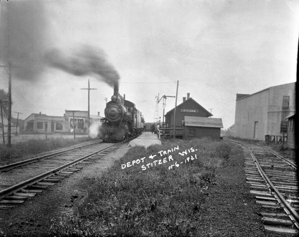 View down side of railroad tracks towards the depot, with a locomotive and passenger cars arriving. People are standing on the platform. In the background are freight cars and a windmill, as well as some storefronts. On the right is a warehouse.