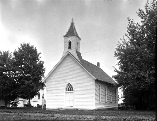 Three-quarter view of front and side of the Methodist Evangelical Church, a wooden building with arched windows on the side and an arched entrance in the front, and a belfry and steeple. A house is next door behind trees.