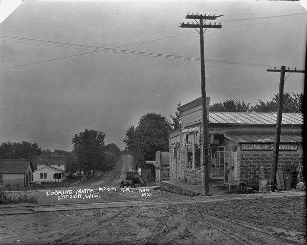 A view down unpaved road of the railroad crossing at Prospect Avenue. A house is down the hill on the left. A hardware store and garage are on the right next to the railroad tracks. An air pump is next to the hardware store. A car is parked in front of the garage. Behind the car there's a highway sign with mileage to Boscobel, Madison and other destinations. Leaning against the hardware store is rolled up fencing, gates, a wagon and a metal barrel.