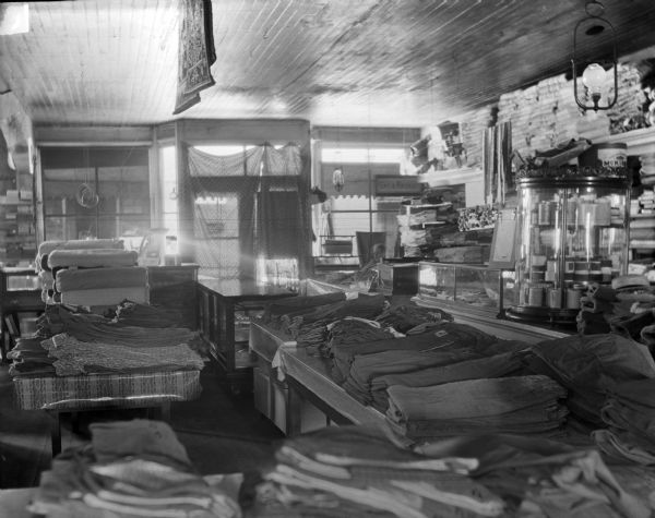 Interior of a clothing store looking towards front of store. In the foreground on tables are piles of folded pants. Bolts of cloth and shirts are on the table on the left. A glass case is near the door, and on the right a woman sits behind the counter. There is a roll of wrapping paper in the window. Two rugs are hanging from the ceiling.