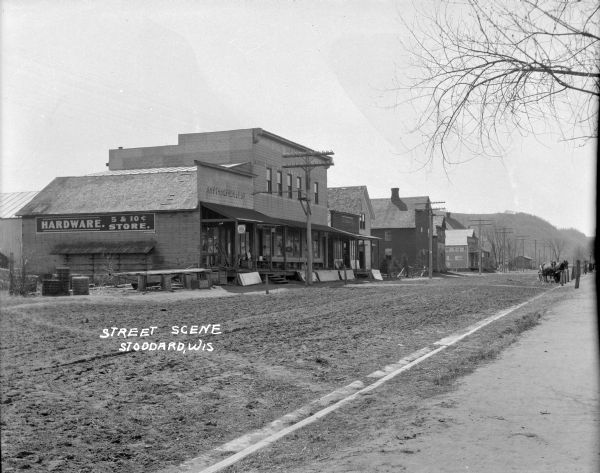 View across street of the central business district. A hardware/5&10 cent store is on the left, with rolled up fencing and various implements on the porch. Louis Place is on the next block. On the right a wagon with two horses is hitched to a post. There are hills in the background.