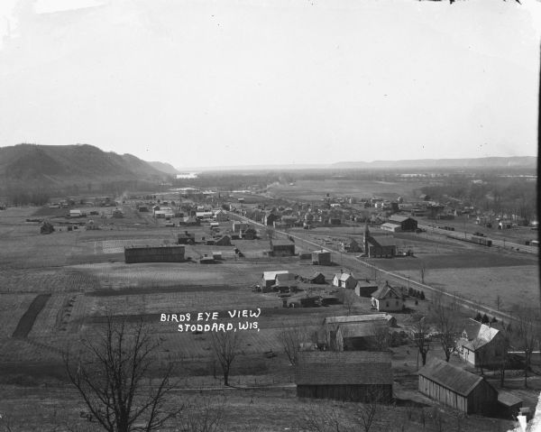 View down hill towards Stoddard, with the Mississippi River and hills in the background. The German Lutheran Church is in the foreground, and there are railroad tracks on the right, with a water tower just past the depot.