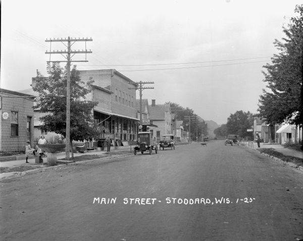 View down Main Street. On the left are two children standing on a stoop near a large drinking fountain. Down the block is a man standing in front of a 5&10 cent store. Next door is an auto supply store and a gas pump, with a delivery truck and an automobile parked in front at the curb. On the right is a restaurant and a man standing near another parked car. In the far background is a tree-lined bluff.