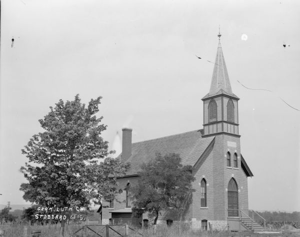 Three-quarter view of front and side of the German Lutheran Church, a brick building with steps to the entrance. There are narrow arched windows, a belfy with shutters, and a steeple. There is a fence in front of the church and two trees. In the background is a cornfield.