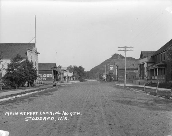 View down center of Main Street. Unidentified buildings are on the left, with a stack of barrels in front. A building with a public phone is next door. Two children are on the sidewalk in front of another storefront. In the distance horses and wagons are hitched up at the curb. On the right is a building with a large porch and a bar next door. A 5&10 cent store is further down the block. In the far distance is a bluff.