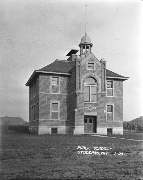 View of front of the public school, a two-story brick building with a cupola and flagpole over the bell. A cornfield and bluff are in the background.