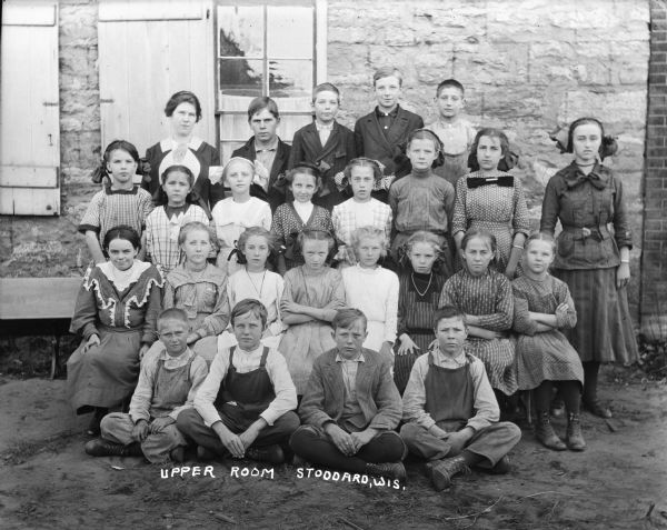 A class picture with four rows posing outdoors, with a stone wall behind them on the left, and a brick wall on the right. In front is a row of boys sitting on the ground. The second row of girls is sitting in chairs, the third row of girls is standing, and in the back, boys are standing with the female teacher. Several of the girls are wearing big hair ribbons.